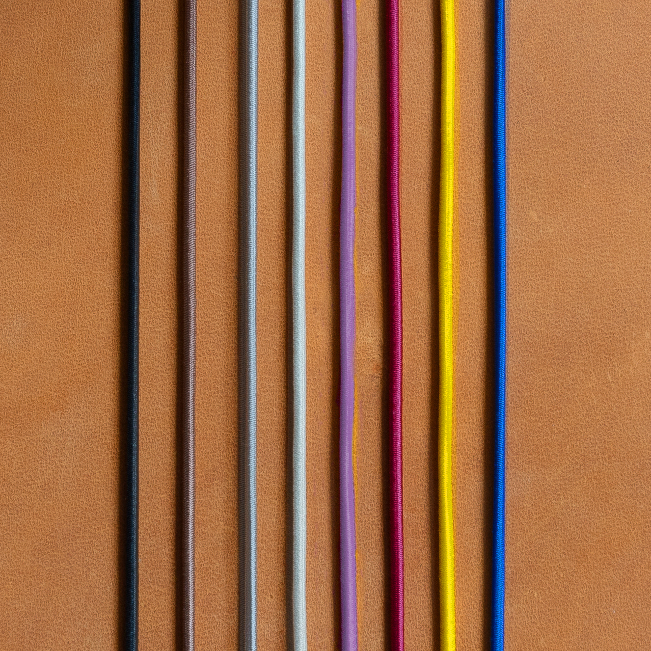 Gummibänder für Notizbuchhüllen in den Farben Schwarz, Braun, Grau, Creme, Rot, Gelb und Blau auf cognacfarbenem Naturleder.