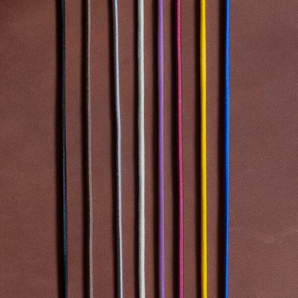 Gummibänder für Notizbuchhüllen in den Farben Schwarz, Braun, Grau, Creme, Rot, Gelb und Blau auf dunkelbraunem Naturleder.