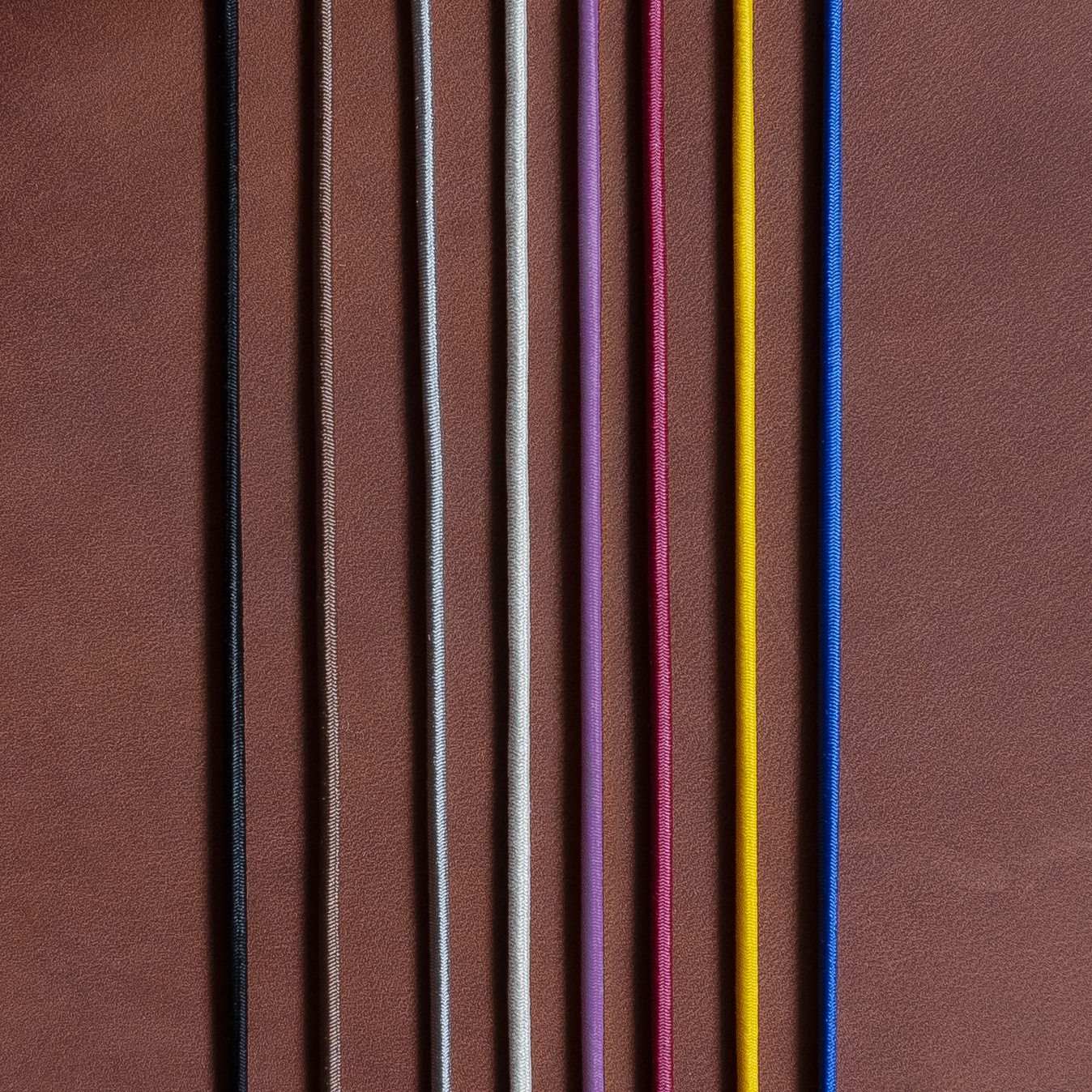 Gummibänder für Notizbuchhüllen in den Farben Schwarz, Braun, Grau, Creme, Rot, Gelb und Blau auf dunkelbraunem Naturleder.