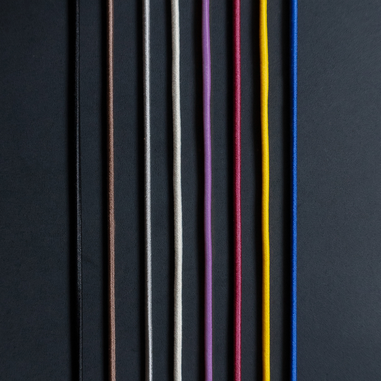 Gummibänder für Notizbuchhüllen in den Farben Schwarz, Braun, Grau, Creme, Rot, Gelb und Blau auf kohlefarbenem Naturleder.