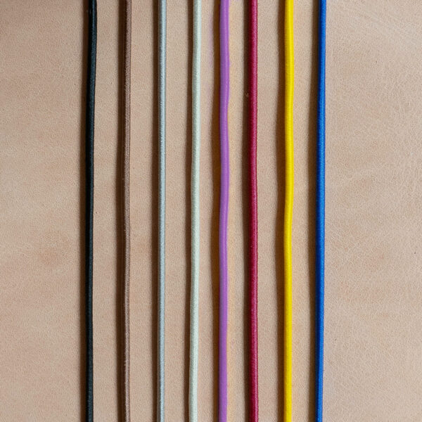 Gummibänder für Notizbuchhüllen in den Farben Schwarz, Braun, Grau, Creme, Rot, Gelb und Blau auf hellbraunem, Naturleder.