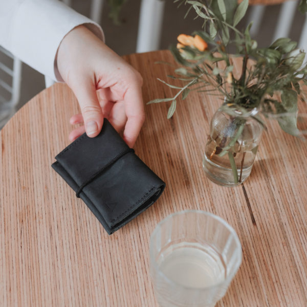 Portemonnaie OLI aus Naturleder in Kohle mit schwarzem Verschlussband auf Holztisch mit Wasserglas