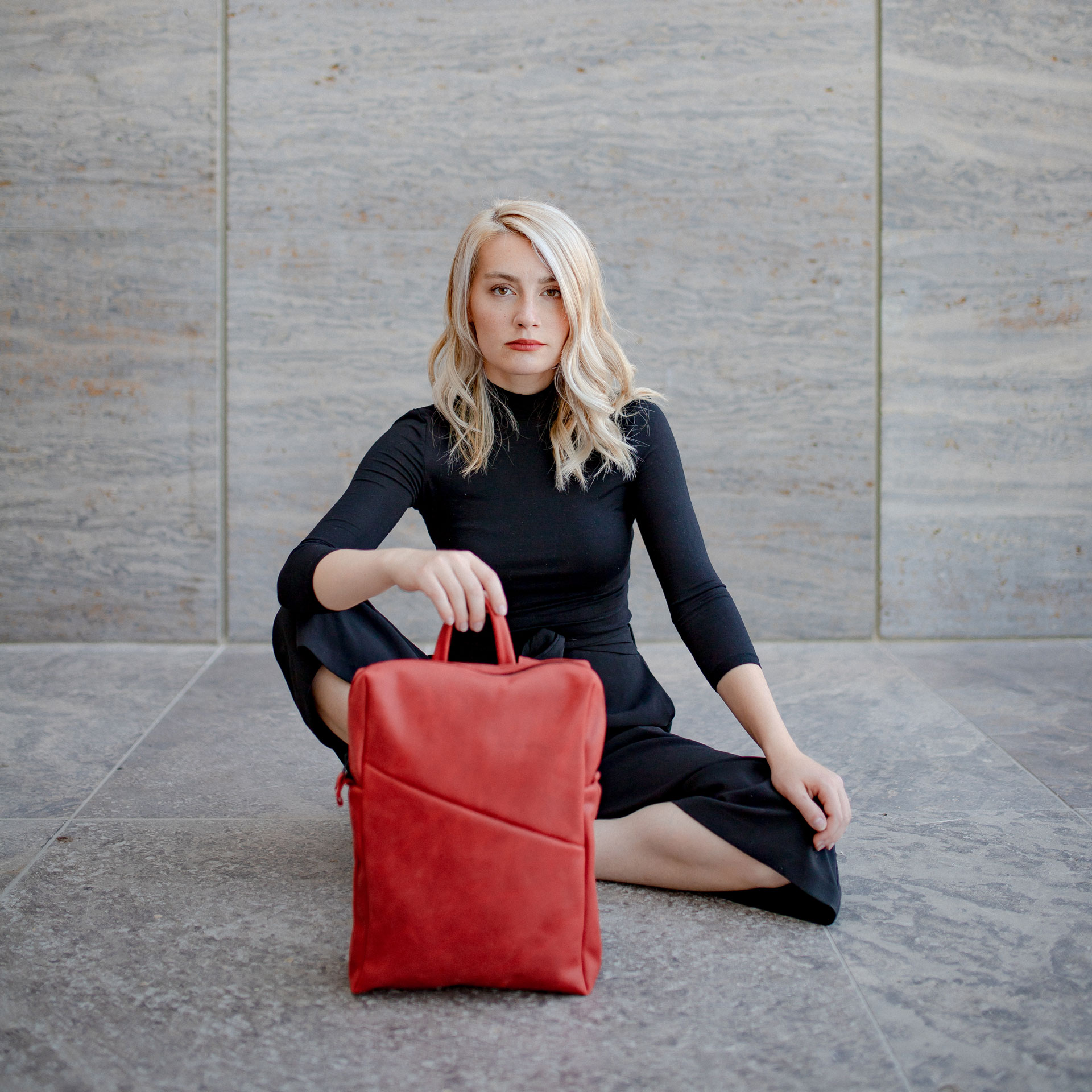 Model sitzt auf dem Boden und hat den Rucksack NEO small in der Farbe Rot vor sich stehen.