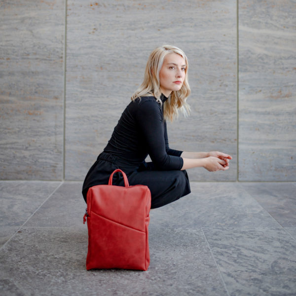 Model hockt auf dem Boden im Seitenprofil und hat den Rucksack Neo small in der Farbe Rot neben sich stehen.