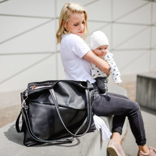 Model sitzt mit ihrem Baby auf dem Schoß da. Vor ihr steht die Wickeltasche MIA Large in der Farbe schwarz geölt mit einer Diagonale in Kohle.