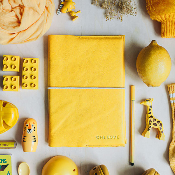 Mood Shot U-Hefthülle EVE aus nachhaltigem Naturleder in Curry mit individueller Prägung in Gold und cremefarbenem Verschlussband neben gelbem Spielzeug